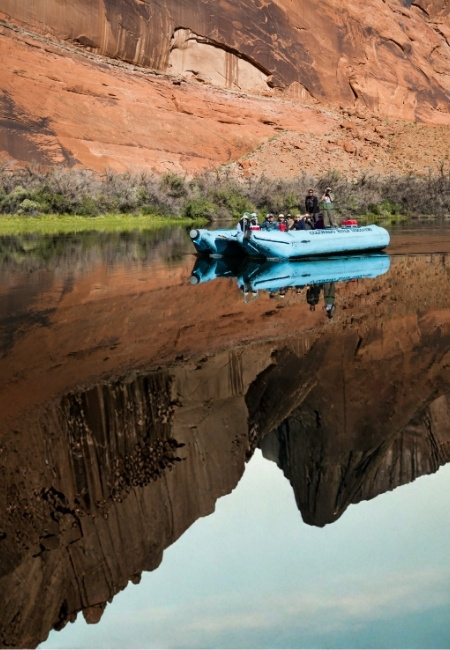 Ein blaues Schlauchboot mit mehreren Personen gleitet auf dem Colorado River vor roten Felswänden und spiegelt sich im ruhigen Wasser des Canyons.