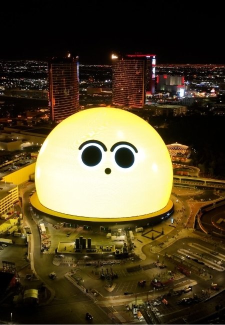 Über dem nächtlichen Las Vegas strahlt die riesige Sphere mit freundlichem Gesicht, während die Hotels am Strip im Hintergrund leuchten.