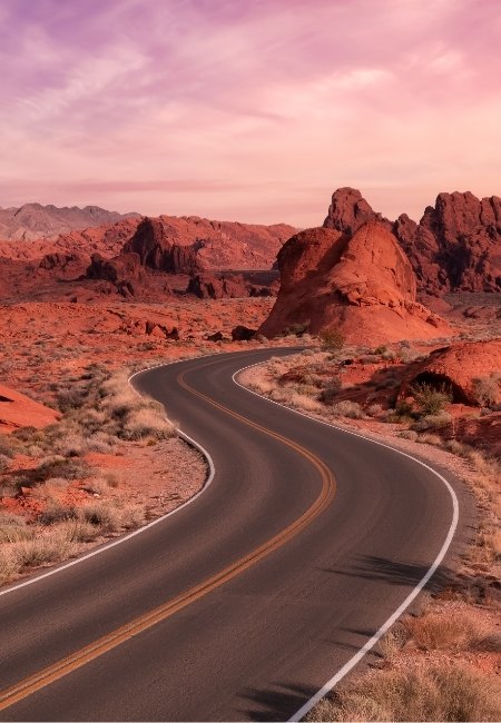 Eine geschwungene Straße führt durch die roten Felsformationen im Valley of Fire nahe Las Vegas unter einem rosa Abendhimmel bei Sonnenuntergang.