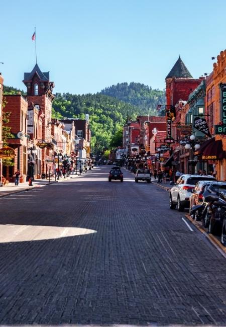 Die Hauptstraße von Deadwood verläuft zwischen historischen Fassaden, geparkten Autos und einem grünen Hügel im Hintergrund.