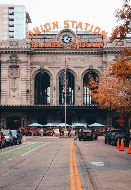 Vor der Denver Union Station ist der Schriftzug „Travel by Train“ zu sehen, dazu herbstliche Bäume und eine belebte Straße.
