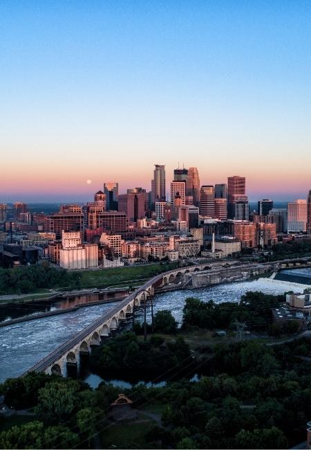 Die Skyline von Minneapolis leuchtet im Abendlicht, während sich eine lange Bogenbrücke über den Mississippi zieht.