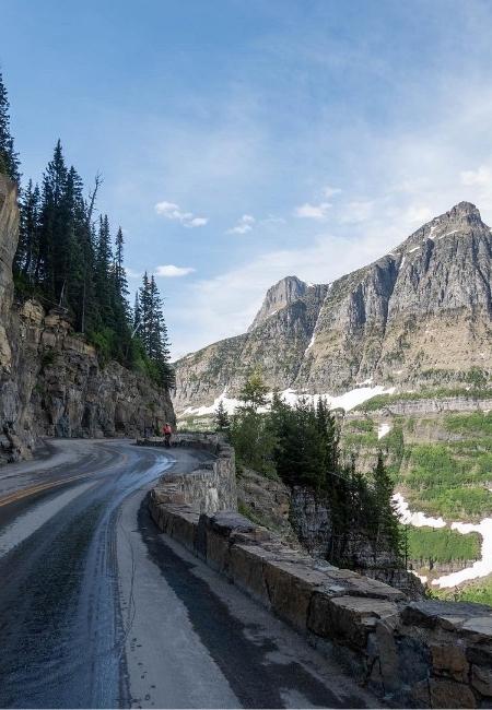 Eine kurvige Bergstraße verläuft an Felswänden entlang, mit Tannen, Schneefeldern und hohen Gipfeln in Montana.