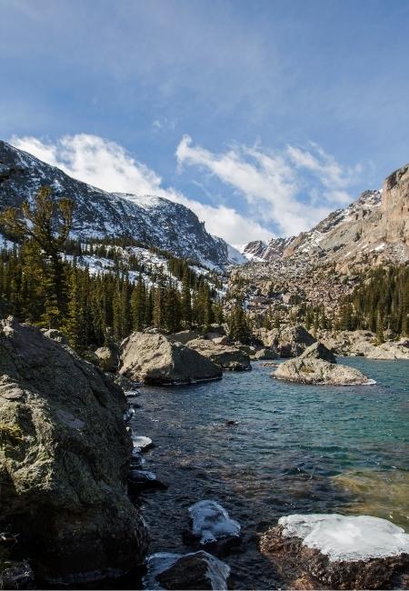 Im Rocky Mountain Nationalpark liegt ein klarer Gebirgssee zwischen Felsen, Wald und schneebedeckten Bergflanken.