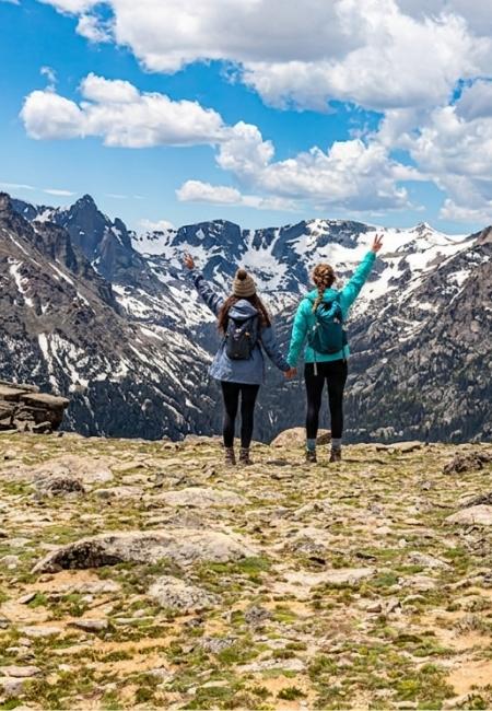 Zwei Wanderinnen stehen auf einem felsigen Hochplateau und blicken auf schneebedeckte Gipfel im Rocky Mountain Nationalpark.