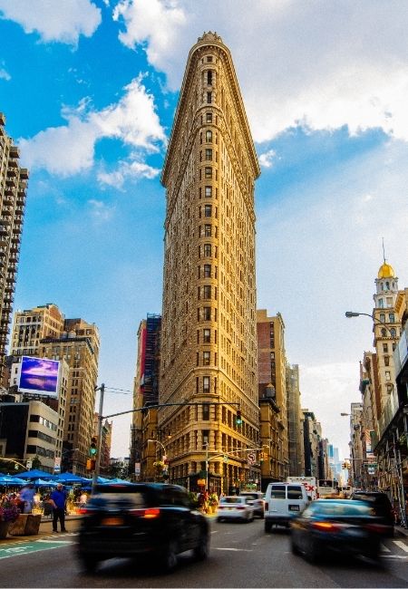 Das Flatiron Building ragt an einer belebten Straßenecke in Manhattan zwischen vorbeifahrenden Autos und hohen Häusern auf.