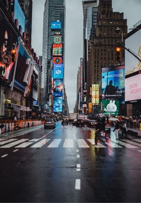 Nasse Fahrbahn und blinkende Reklametafeln prägen die geschäftige Szenerie am Times Square in Manhattan, New York.