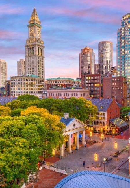 Die Skyline von Boston zeigt den historischen Turm am Hafenviertel vor modernen Hochhäusern und grünen Bäumen im Vordergrund.