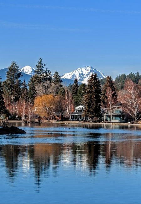 Vor ruhigem Wasser spiegelt sich eine Uferlinie mit Häusern und Bäumen, dahinter ragen schneebedeckte Berge in den blauen Himmel.