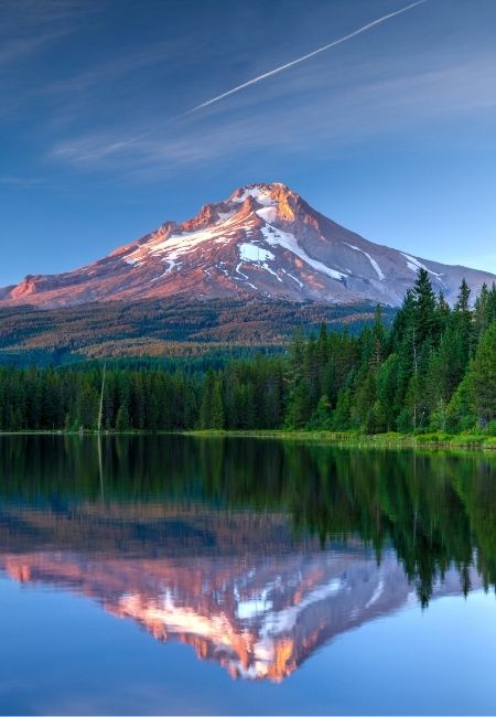 Mount Hood ragt schneebedeckt über einen stillen Bergsee, während sich der Gipfel im klaren Wasser und am Waldsaum spiegelt.