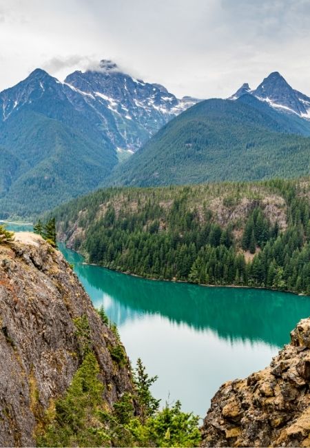 Im North Cascades National Park liegt ein türkisfarbener See vor bewaldeten Hängen und schneebedeckten Bergen.