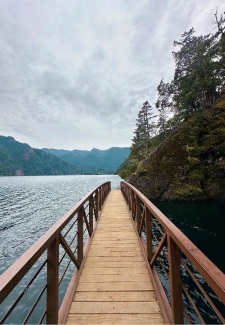 Ein schmaler Holzsteg führt entlang der felsigen Küste bei Port Angeles über ruhiges Wasser und an bewaldeten Hängen vorbei.