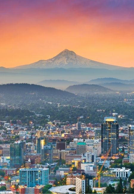 Portland zeigt sich unter einem orangefarbenen Abendhimmel, während Mount Hood schneebedeckt über der Stadt aufragt.