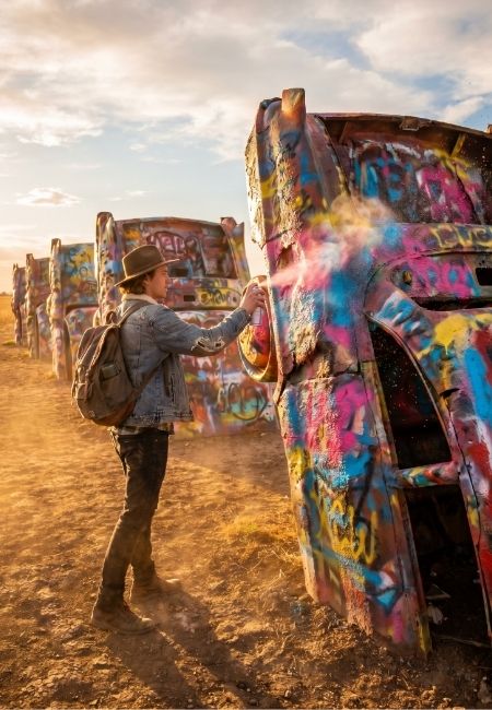 Eine Person besprüht ein bunt graffiti-bedecktes Auto an der Cadillac Ranch entlang der Route 66 im warmen Abendlicht.