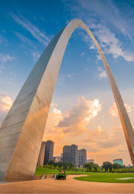 Der Gateway Arch in St. Louis ragt vor einem warmen Abendhimmel über die Grünanlagen und die Skyline am Mississippi auf.