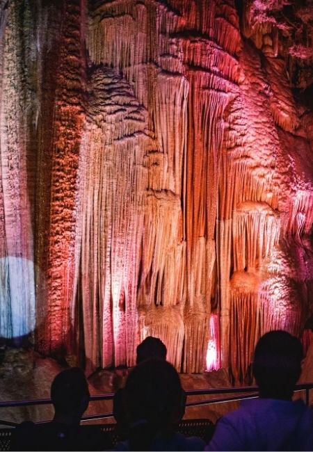 In den Meramec Caverns leuchtet eine hohe Tropfsteinwand in Rot- und Violetttönen, davor sitzen Besucher.