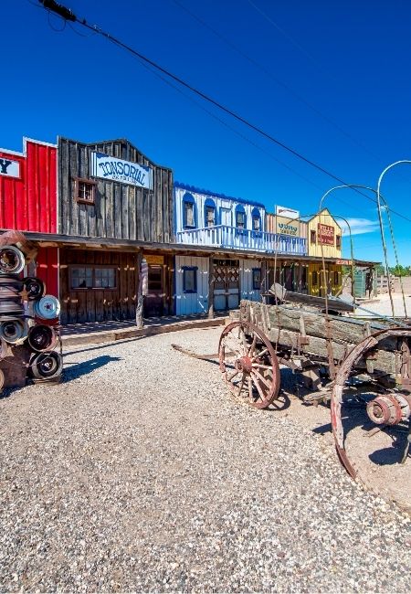 Bunte Holzhäuser mit Western-Charakter und ein alter Wagen stehen auf kiesigem Boden unter tiefblauem Himmel an der Route 66.