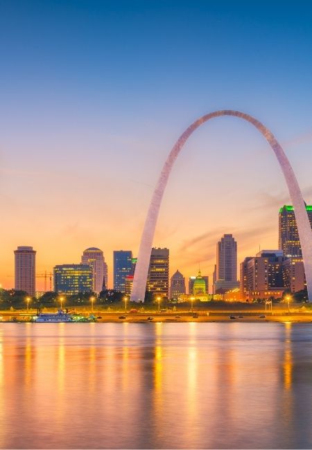 Über der Skyline von St. Louis spannt sich der Gateway Arch bei Sonnenuntergang, und die Lichter spiegeln sich im Fluss.
