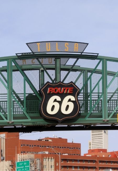 Die grüne Brücke in Tulsa trägt den markanten Route-66-Schriftzug vor der Stadtkulisse und dem hellen Himmel.