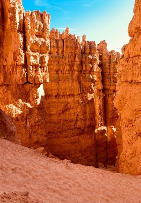 Im Bryce Canyon ragen orangefarbene Felsnadeln und steile Wände über einem sandigen Canyonboden in den blauen Himmel.