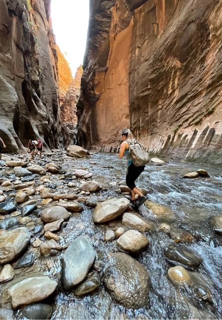 Eine Person watet mit Rucksack durch den flachen Fluss zwischen hohen Felswänden in den Narrows des Zion-Nationalparks.