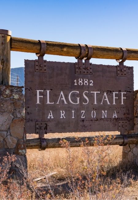 Ein rustikales Holzschild mit der Aufschrift „Flagstaff Arizona“ steht vor trockenem Gras und Himmel.
