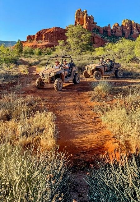 Zwei offene Jeeps fahren auf einem staubigen Wüstenpfad vor den roten Felsen von Sedona durch karges Buschland.