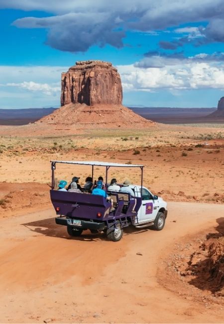 Ein offener Geländewagen mit Tourgästen fährt auf einer staubigen Piste vor einem mächtigen Tafelberg in Monument Valley.