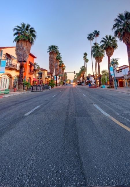 Eine breite Straße in Palm Springs ist von hohen Palmen und farbigen Gebäuden gesäumt, im Abendlicht unter klarem Himmel.