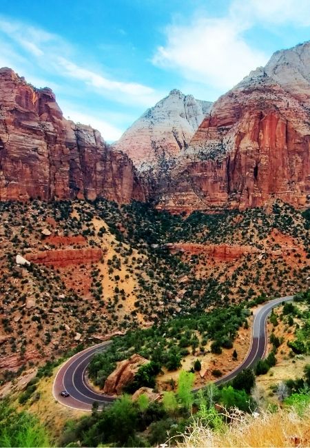 Vor hohen roten Felswänden im Zion National Park schlängelt sich eine Serpentinenstraße durch die trockene Canyonlandschaft.