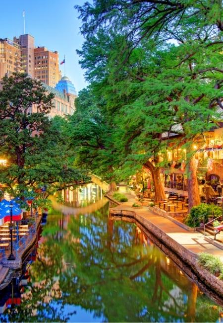 Der River Walk in San Antonio zeigt einen beleuchteten Kanal mit Restaurants, großen Bäumen und Spiegelungen im Wasser.
