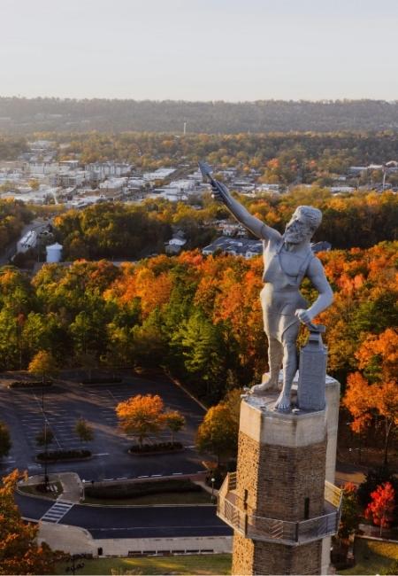 Die Vulcan-Statue in Birmingham, Alabama ragt über bunte Herbstwälder und die Stadt im Abendlicht.
