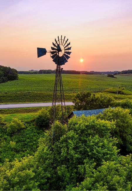 Eine hohe Windmühle steht über grünen Feldern im Texas Hill Country, während die tief stehende Sonne den Himmel orange färbt.