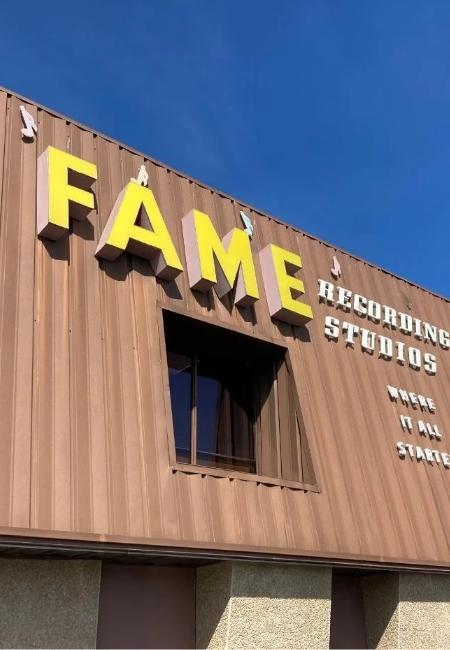 Am Gebäude der FAME Recording Studios in Muscle Shoals leuchten gelbe Buchstaben vor brauner Fassade unter blauem Himmel.
