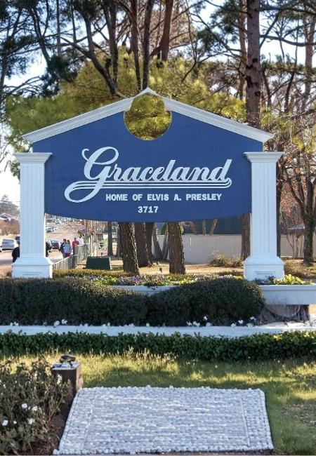 Vor dem blauen Graceland-Schild mit der Aufschrift Home of Elvis A. Presley 3717 stehen Bäume und Sträucher.