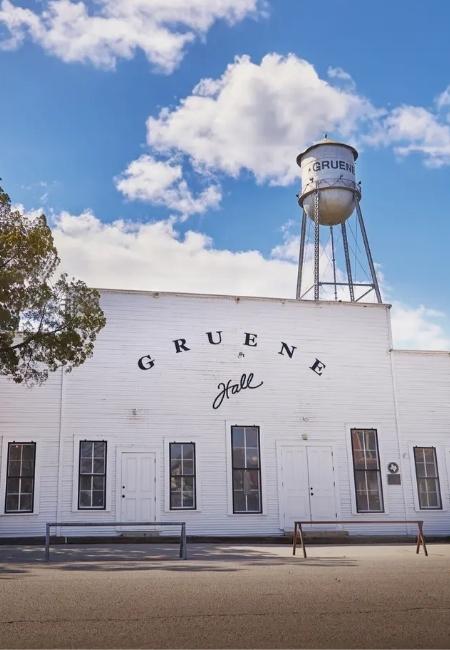 Die weiße Gruene Hall in New Braunfels, Texas steht unter einem Wasserturm vor blauem Himmel mit lockeren Wolken.