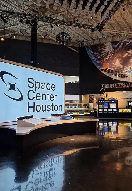 Im Space Center Houston steht eine große Empfangswand neben interaktiven Ausstellungsinseln unter dem Hallendach.