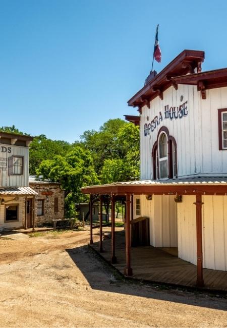 Das weiße Opera House mit rotem Dach steht in Wimberley, Texas, neben weiteren historischen Gebäuden unter blauem Himmel.
