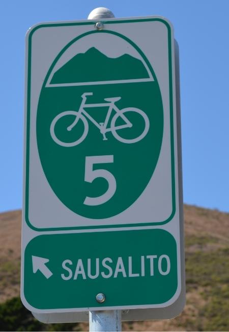 Ein grünes Wegschild zeigt die Fahrradroute 5 nach Sausalito mit Pfeil und Fahrradsymbol vor blauem Himmel.