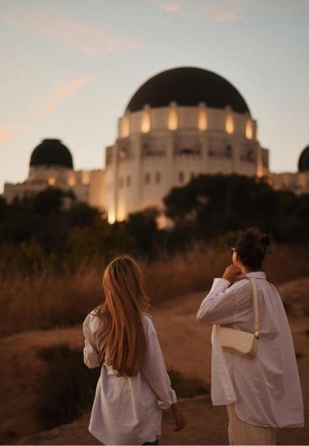 Zwei Frauen gehen auf einem Pfad zum Griffith Observatory, dessen Kuppeln im Abendlicht über Los Angeles leuchten.