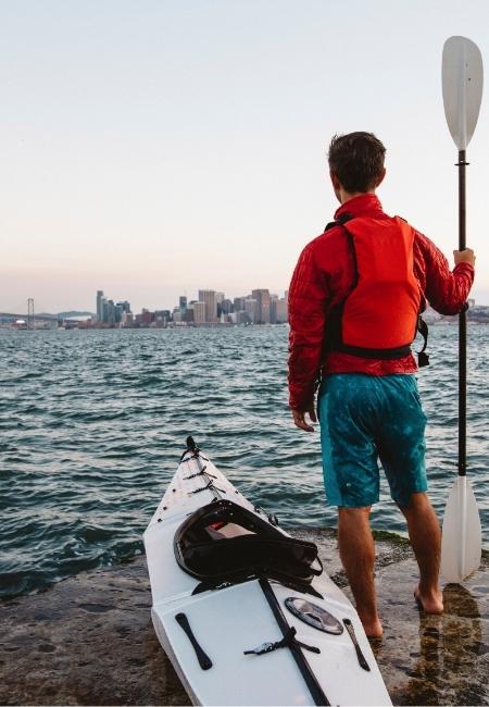 Ein Kajakfahrer mit roter Schwimmweste steht am Ufer der Monterey Bay und blickt auf die Skyline am Wasser.
