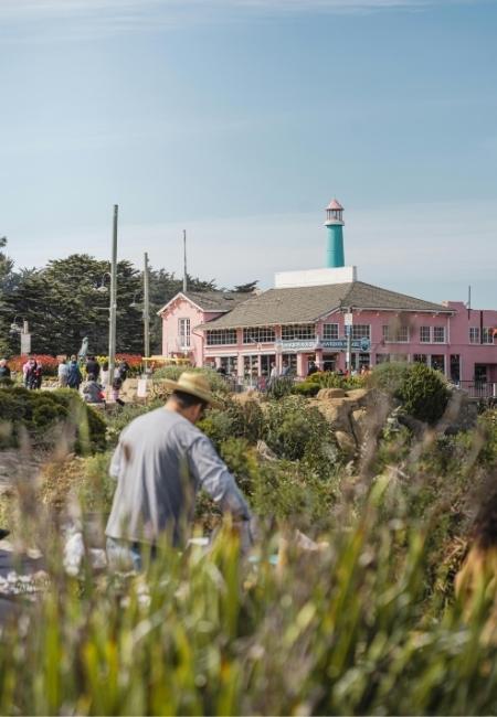 Vor einem rosa Gebäude in Monterey steht ein türkisfarbener Turm, davor Gras und ein Mann mit Hut.