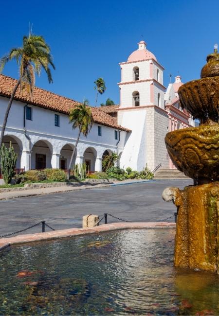 Vor dem weißen Missionsbau in Santa Barbara stehen Palmen und ein Brunnen, während der wolkenlose Himmel die Szene prägt.