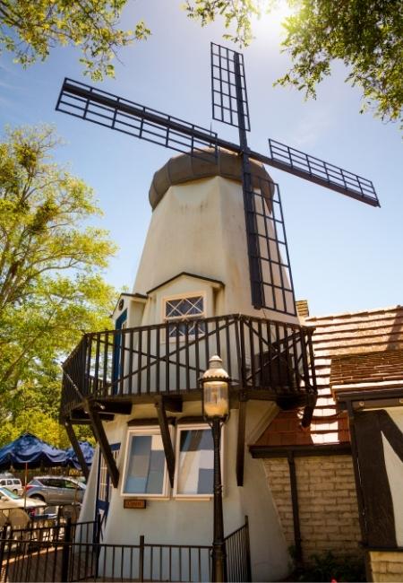 In Solvang steht eine hohe Windmühle mit schwarzem Flügelrad neben einem Fachwerkhaus und einem kleinen Balkon.
