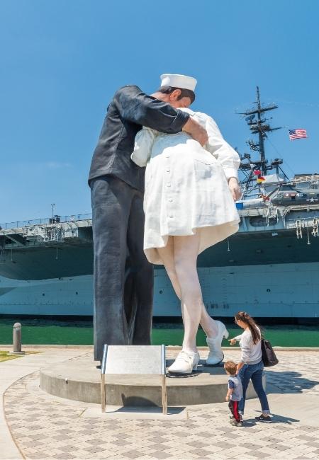 Vor der USS Midway in San Diego steht ein großes Matrosenpaar, daneben schaut eine kleine Familie hin.