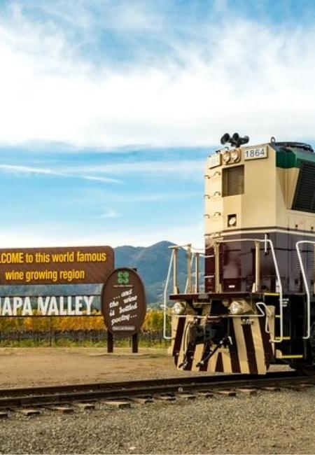 Vor dem Schild zum Napa Valley steht eine Lokomotive am Gleis, dahinter liegen Weinberge und Hügel unter blauem Himmel.