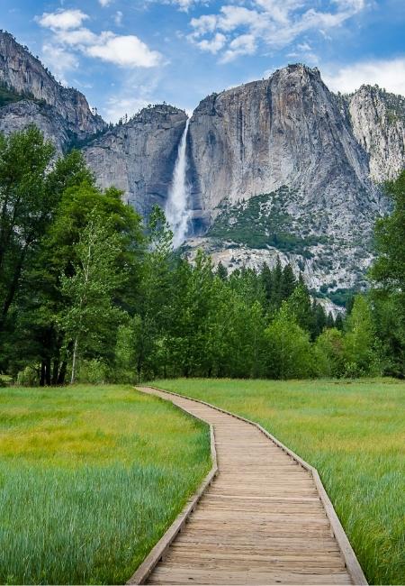 Ein Holzsteg führt durch eine grüne Wiese im Yosemite Nationalpark auf einen hohen Wasserfall vor steilen Granitwänden zu.