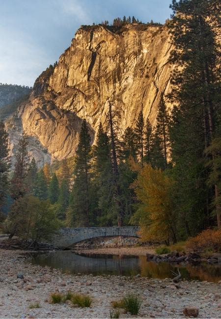 Die goldene Granitwand im Yosemite Valley ragt über Kiefern, Fluss und eine kleine Steinbrücke im warmen Abendlicht auf.