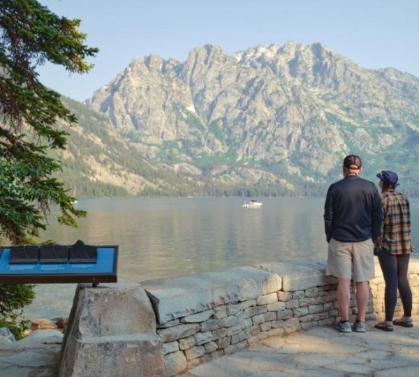 Zwei Besucher blicken am Seeufer auf die markante Bergkulisse des Grand Teton National Park in Wyoming.