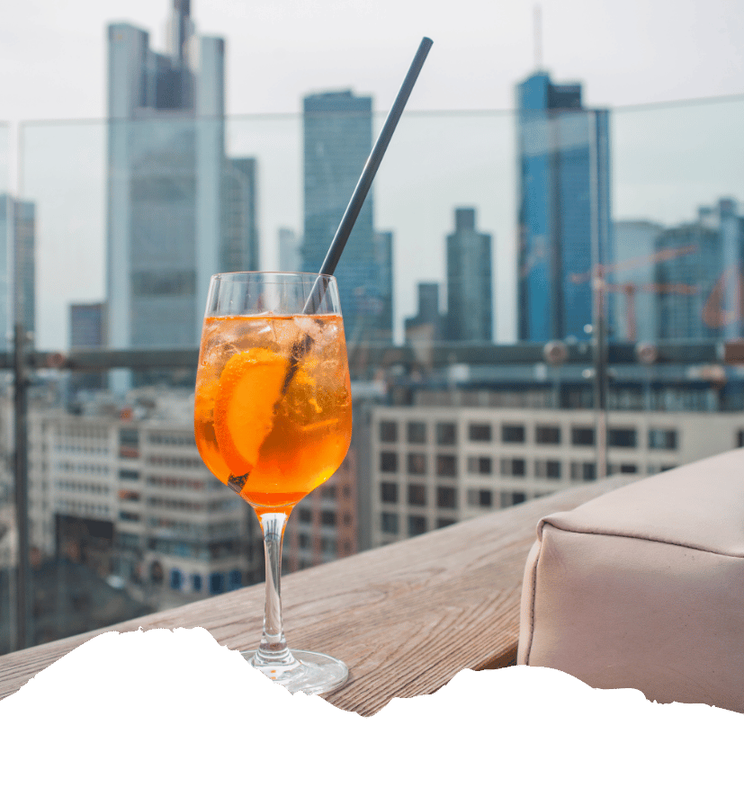 Ein orangefarbener Drink mit Orangenscheibe steht auf einer Dachterrasse vor der Skyline von New York.